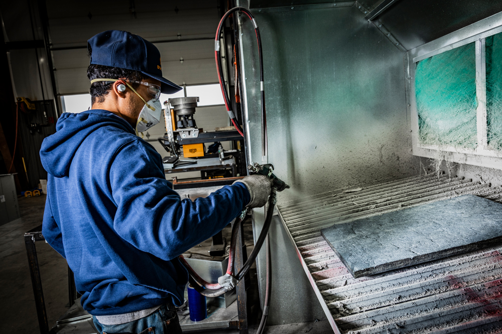 A DuctWorx technician applying adhesive coating to duct liner at a spray booth, preparing sound-attenuated panels for lined rectangular duct systems.